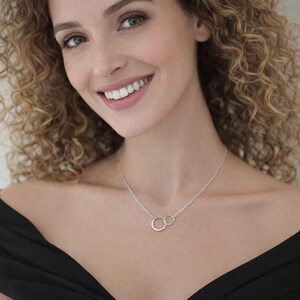 May include: A silver necklace featuring two interlocking circular rings as a pendant on a delicate chain. The necklace rests against the collarbone, showcasing a polished metallic finish and a simple, clean design that connects two distinct circles.