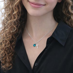 May include: A silver necklace with a cross pendant and a teal-colored gemstone charm. The necklace is worn against a black shirt, creating a contrast that highlights the jewelry's details. The cross is a simple, elegant design.