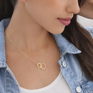 May include: A gold-toned necklace featuring two interlocking circular rings as a pendant, suspended from a delicate, thin chain. The necklace rests against the neck, showcasing a simple, polished metal finish on both the rings and the chain links.