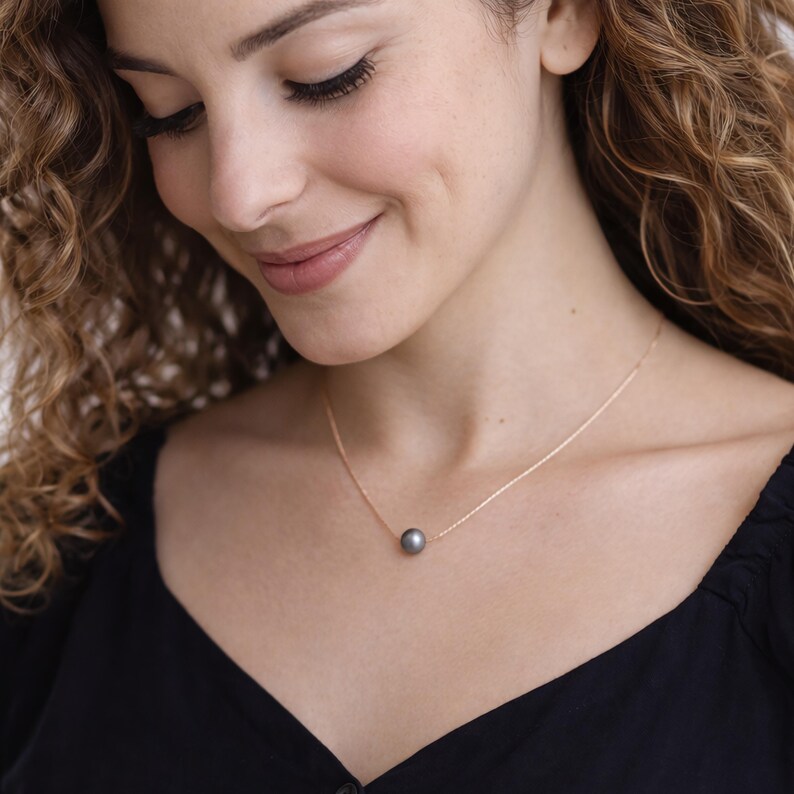 Puede incluir: Un delicado collar de oro con un colgante de perla gris oscuro. El collar se lleva sobre una blusa negra, resaltando el contraste entre la perla y la cadena de oro. La mujer de la imagen est&aacute; sonriendo.