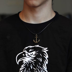 May include: A person wearing a black t-shirt with a white eagle graphic and a gold anchor necklace.