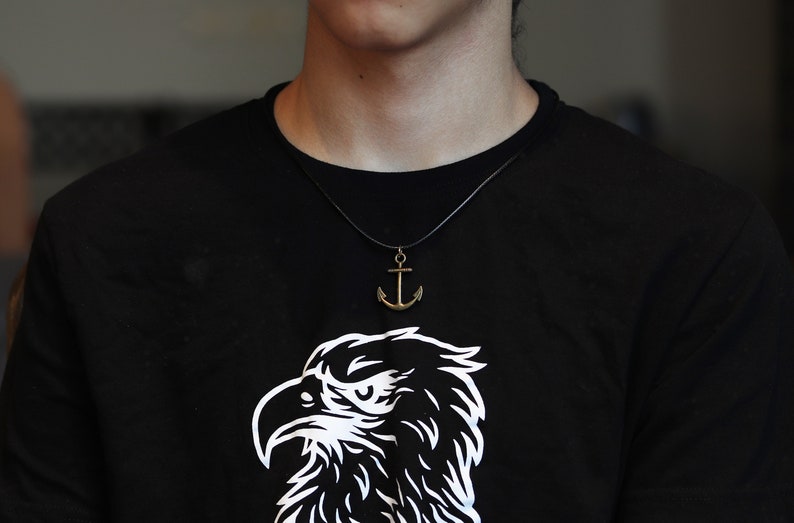 May include: A person wearing a black t-shirt with a white eagle graphic and a gold anchor necklace.
