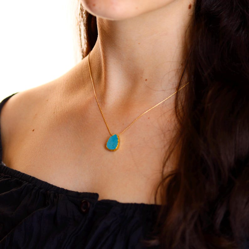May include: A gold necklace featuring a teardrop-shaped pendant with a vibrant turquoise stone. The pendant is outlined in gold, and the necklace is worn on a person's neck, against a dark top.