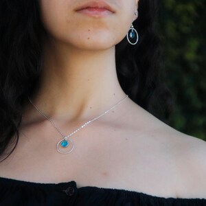 Puede incluir: Un collar de plata con un colgante circular que presenta una peque&ntilde;a piedra preciosa redonda de color azul brillante en el centro, junto con pendientes de aro de plata a juego, cada uno con una piedra azul redonda id&eacute;ntica suspendida en el marco.