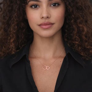 May include: A rose gold necklace featuring two interlocking circular pendants suspended from a delicate chain. The metal has a smooth, polished finish, and the two rings are connected in a side-by-side arrangement, resting against the collarbone.