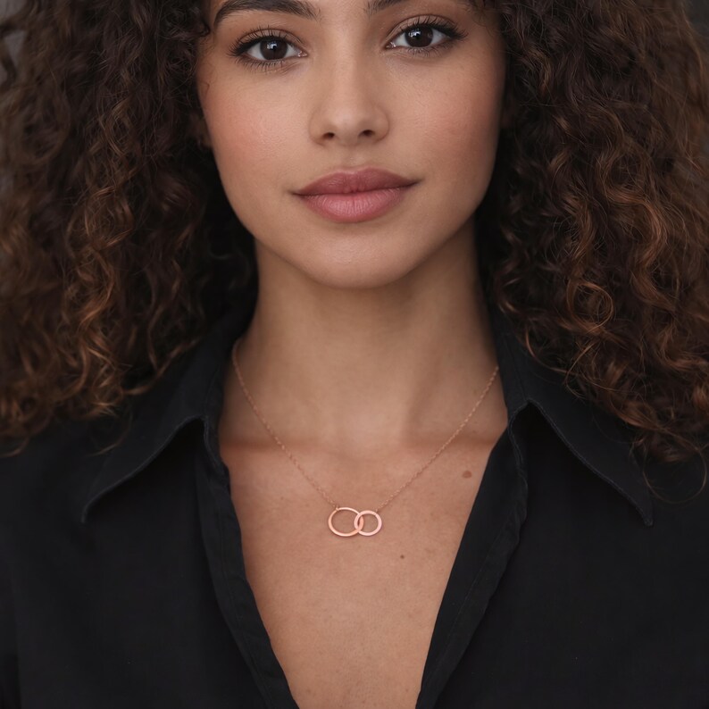 May include: A rose gold necklace featuring two interlocking circular pendants suspended from a delicate chain. The metal has a smooth, polished finish, and the two rings are connected in a side-by-side arrangement, resting against the collarbone.