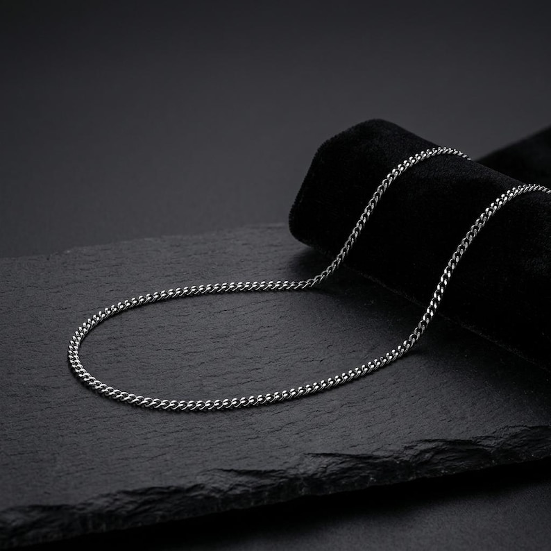 May include: A silver-toned metal chain necklace featuring a classic curb link design. The chain is composed of small, uniform, interlocking oval links that create a smooth, continuous, and flexible strand with a polished, reflective metallic finish.