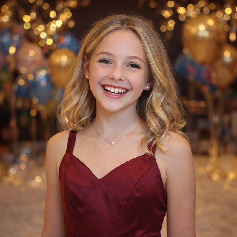 May include: A smiling young person with blonde hair and a burgundy dress. They are wearing a delicate necklace with a small pendant. The background features blurred balloons and lights, creating a festive atmosphere.