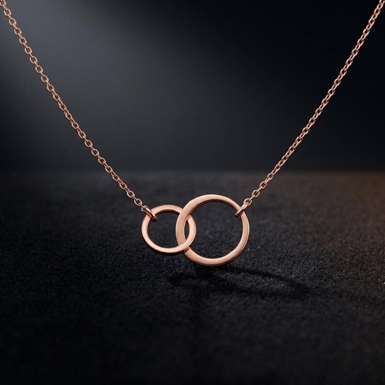 May include: A rose gold necklace featuring two interlocking circular rings of different sizes suspended from a delicate chain. The metal has a polished finish, and the design consists of a smaller ring linked to a larger ring, creating a continuous loop.