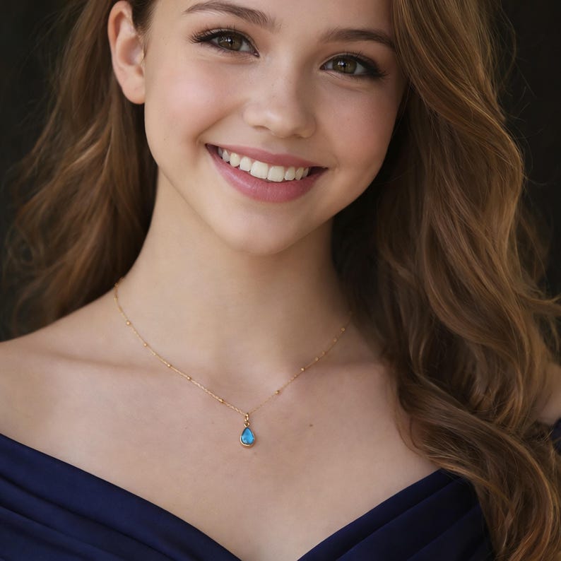 Puede incluir: Un collar dorado con un peque&ntilde;o colgante azul claro en forma de l&aacute;grima. La cadena tiene peque&ntilde;as cuentas doradas. La mujer de la imagen lleva un vestido azul oscuro.