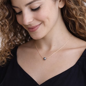May include: A delicate gold necklace featuring a single, dark gray pearl pendant. The necklace is worn against a black top, highlighting the contrast between the pearl and the gold chain. The woman in the image is smiling.