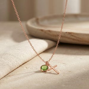 May include: A rose gold chain necklace featuring two charms: a round, faceted, light green peridot gemstone set in a bezel and a simple, plain rose gold cross pendant. The delicate metal chain connects to both charms, which hang together at the center.