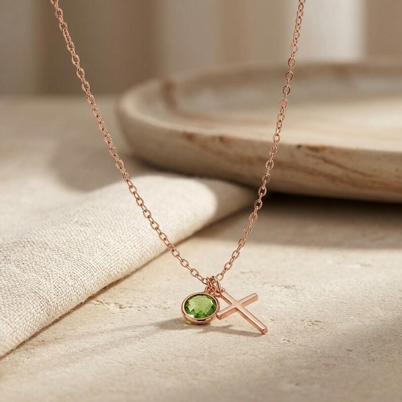 May include: A rose gold chain necklace featuring two charms: a round, faceted, light green peridot gemstone set in a bezel and a simple, plain rose gold cross pendant. The delicate metal chain connects to both charms, which hang together at the center.