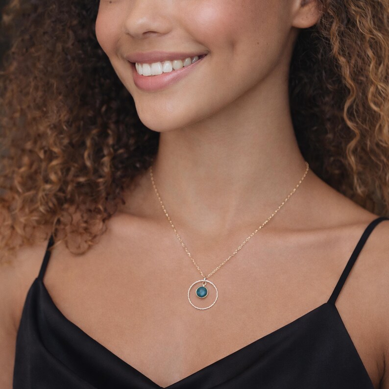 Puede incluir: Un collar de tono dorado con una cadena fina y un colgante circular. El colgante consiste en un anillo fino y texturizado de tono dorado que enmarca una peque&ntilde;a piedra preciosa redonda de color azul verdoso oscuro en el centro.