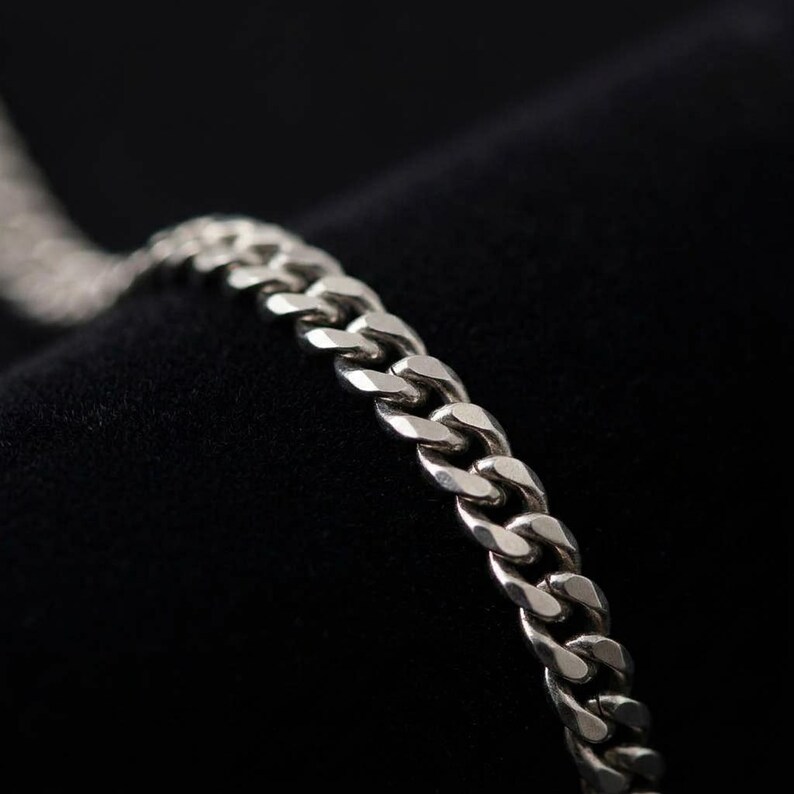 May include: A silver-toned metal chain bracelet featuring a classic curb link design. The individual links are polished and interconnected, creating a continuous, flexible band with a reflective surface that catches the light along its length.
