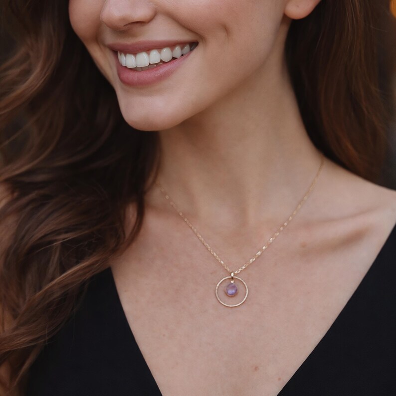 Puede incluir: Un collar de tono dorado con una cadena delicada y un colgante circular. El colgante consiste en un anillo fino de tono dorado que enmarca una peque&ntilde;a piedra preciosa redonda de color morado claro, sujeta por una peque&ntilde;a argolla dorada.