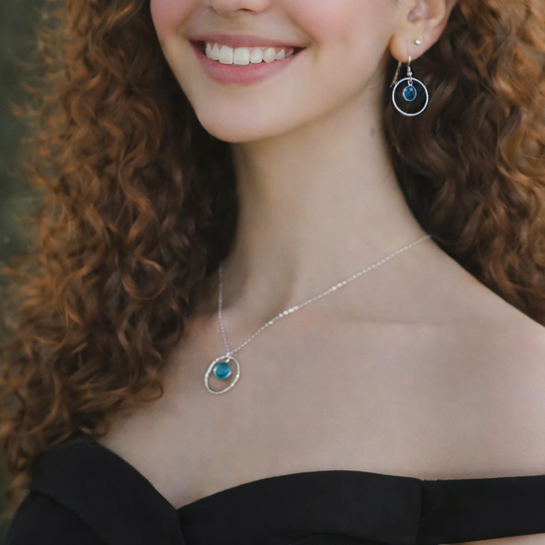 May include: A silver necklace and matching drop earrings, each featuring a circular silver frame holding a small, round, translucent blue stone. The necklace hangs on a delicate silver chain, and the earrings are suspended from silver hooks.