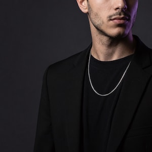 May include: A thin, silver-toned curb chain necklace worn around the neck. The necklace features a series of interlocking, uniform metal links that create a smooth, continuous loop resting against a plain black t-shirt and a black blazer.