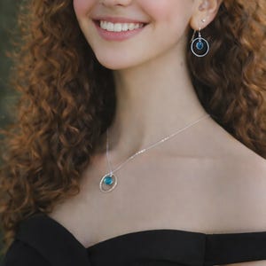 Puede incluir: Un collar y pendientes colgantes a juego en plata, cada uno con un marco circular de plata que sostiene una peque&ntilde;a piedra azul redonda y transl&uacute;cida. El collar cuelga de una fina cadena de plata y los pendientes tienen ganchos de plata.