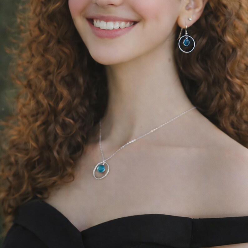 Puede incluir: Un collar y pendientes colgantes a juego en plata, cada uno con un marco circular de plata que sostiene una peque&ntilde;a piedra azul redonda y transl&uacute;cida. El collar cuelga de una fina cadena de plata y los pendientes tienen ganchos de plata.