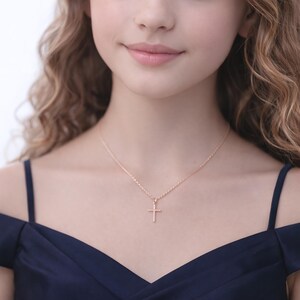 May include: A rose gold cross pendant necklace. The cross is a simple, classic design, suspended from a delicate chain. The necklace is worn against a dark blue dress.