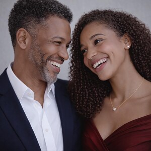 May include: A smiling couple is shown in a close-up shot. The man wears a navy blue suit jacket over a white dress shirt. The woman wears a burgundy dress and a gold necklace with a pearl pendant. Both are looking at each other and smiling.