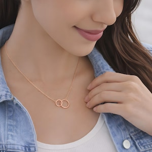 May include: A rose gold necklace featuring two interlocking circular rings as a pendant. The delicate chain is secured around the neck, showcasing a polished metallic finish that catches the light. The design consists of one smaller ring linked to a larger ring.