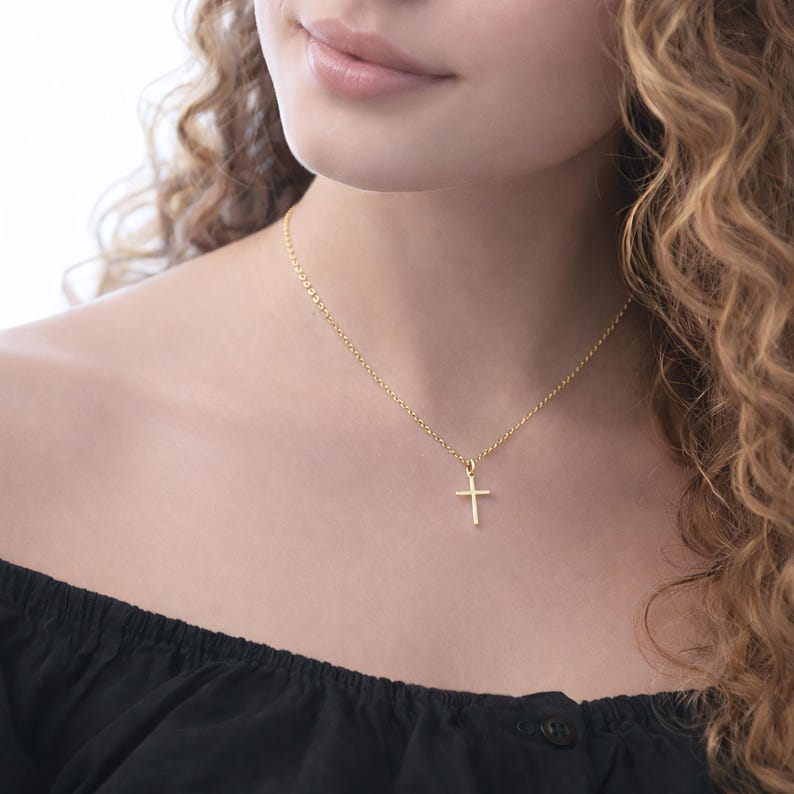 Puede incluir: Un collar dorado con un peque&ntilde;o colgante de cruz. La cadena es fina y la cruz tiene un dise&ntilde;o sencillo y minimalista. El collar se lleva alrededor del cuello. El fondo est&aacute; borroso.