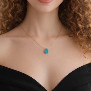 May include: A gold-colored necklace with a teardrop-shaped, turquoise-colored pendant. The pendant has a gold-colored border. The necklace is worn on a person with curly hair and a black top.