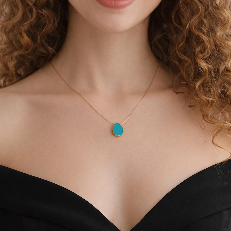 May include: A gold-colored necklace with a teardrop-shaped, turquoise-colored pendant. The pendant has a gold-colored border. The necklace is worn on a person with curly hair and a black top.