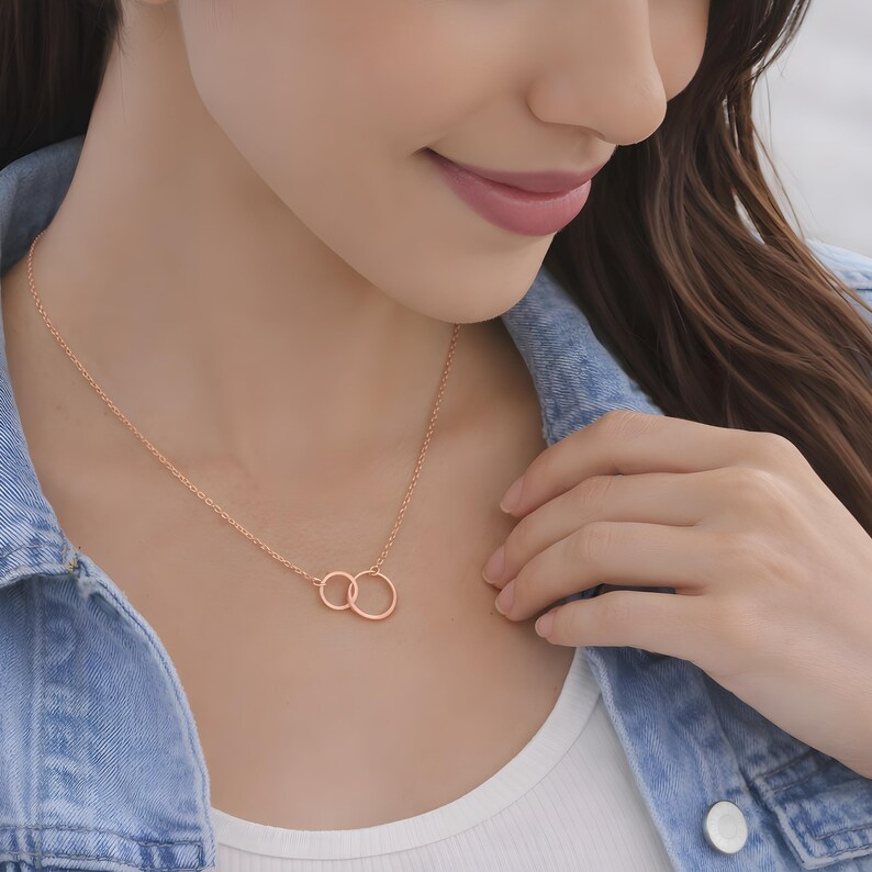 May include: A rose gold necklace featuring two interlocking circular rings as a pendant. The delicate chain is secured around the neck, showcasing a polished metallic finish that catches the light. The design consists of one smaller ring linked to a larger ring.
