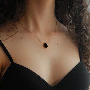 May include: A gold-toned necklace featuring a teardrop-shaped black pendant. The pendant is outlined with a gold-colored metal. The necklace is worn on a person with dark curly hair and a black top.