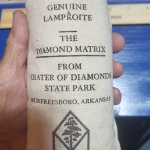 May include: A beige canvas bag with the text "GENUINE LAMPROITE" and "THE DIAMOND MATRIX FROM CRATER OF DIAMONDS STATE PARK MURFREESBORO, ARKANSAS". The bag also features the Arkansas State Parks logo.
