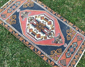 Op de afbeelding: Rechthoekig tapijt met een centraal ruitpatroon in rood, wit en bruin, omlijst door een marineblauwe rand. Het tapijt heeft geometrische ontwerpen in oranje, bruin en blauw, met een gefranjerde rand. Het tapijt ligt op groen gras.