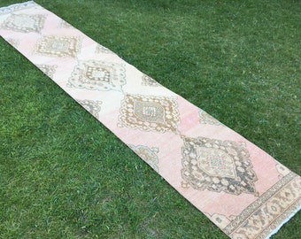 May include: A long, pink and cream runner rug with a geometric pattern. The rug has a fringe border and is laid out on a green lawn.