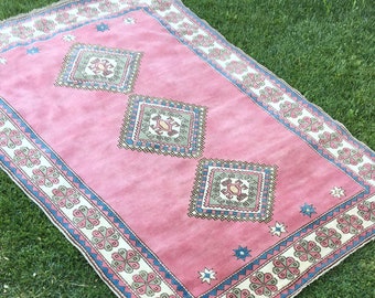 Puede incluir: Una alfombra rosa con un patrón geométrico en tonos de azul, marrón y blanco. La alfombra tiene una forma de diamante en el centro con un diamante más pequeño en su interior. La alfombra está sobre una superficie de hierba verde.