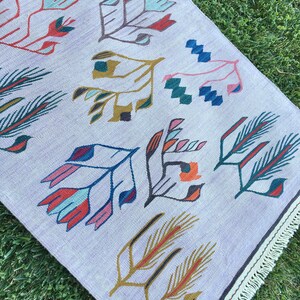 May include: A light purple rug with a geometric pattern of stylized flowers and leaves in shades of red, blue, green, and yellow. The rug has a white fringe border.