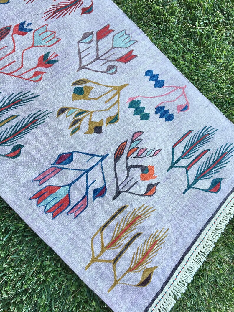 May include: A light purple rug with a geometric pattern of stylized flowers and leaves in shades of red, blue, green, and yellow. The rug has a white fringe border.