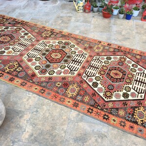 May include: A traditional Turkish rug with a geometric pattern in shades of red, orange, brown, and white. The rug has a central medallion with a hexagonal design and a border with a repeating star pattern.