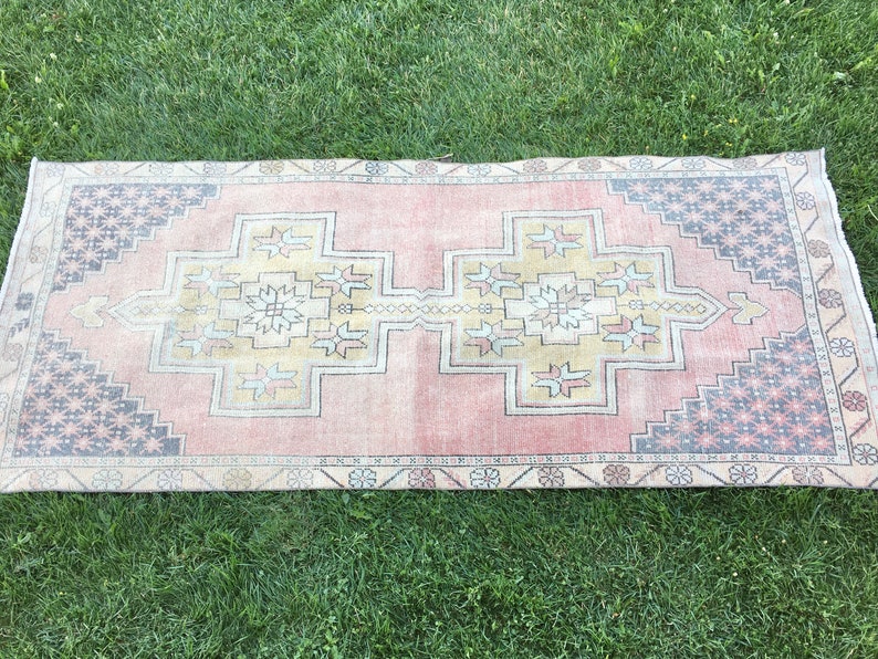 May include: Rectangular rug with a faded pink central field and a geometric pattern in the center. The rug has a border with a repeating star and floral design in shades of gray, blue, and red. The rug is placed on green grass.
