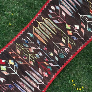 Puede incluir: Una alfombra larga y estrecha con fondo marr&oacute;n y un patr&oacute;n geom&eacute;trico colorido de flores y hojas estilizadas. La alfombra tiene un borde rojo.