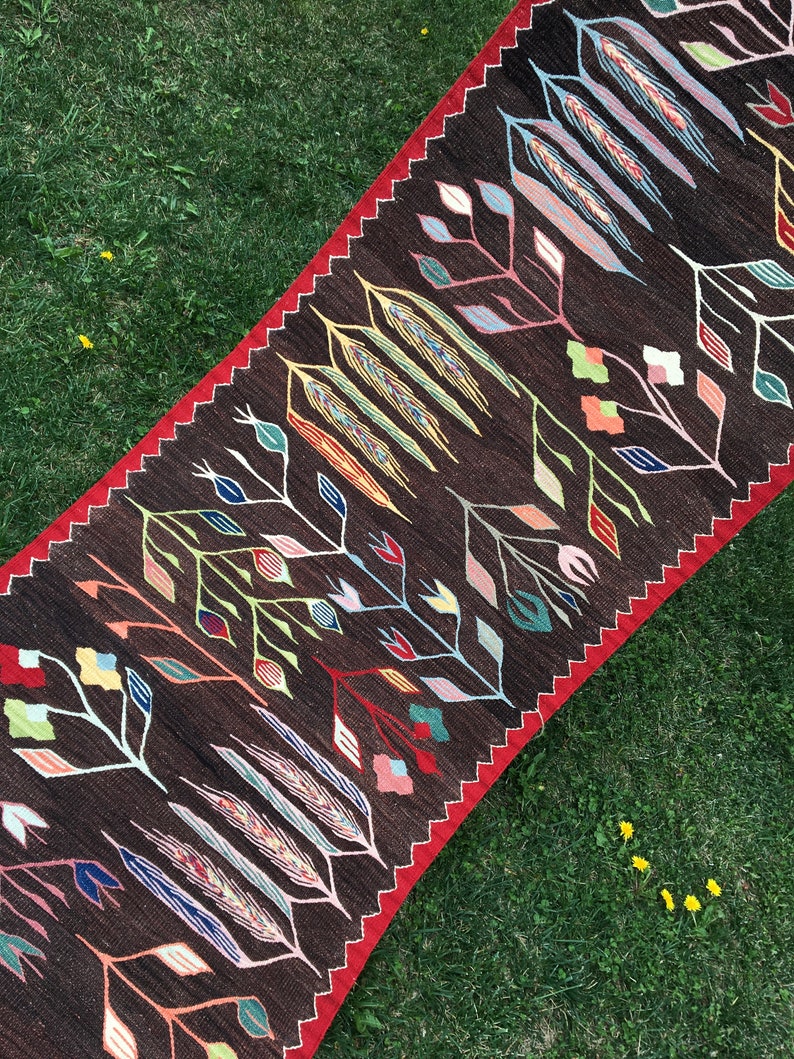 Puede incluir: Una alfombra larga y estrecha con fondo marr&oacute;n y un patr&oacute;n geom&eacute;trico colorido de flores y hojas estilizadas. La alfombra tiene un borde rojo.