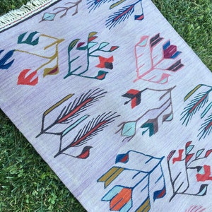 May include: A light purple rug with a geometric pattern of stylized flowers and leaves in shades of blue, red, green, and yellow. The rug has a fringe border.