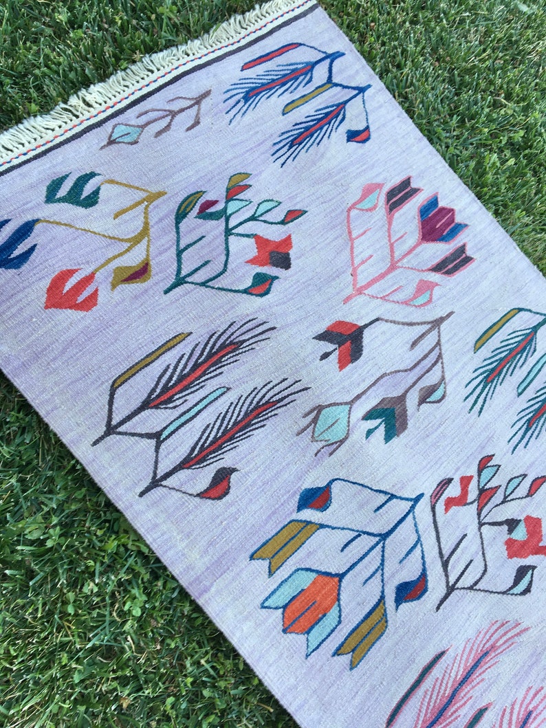 May include: A light purple rug with a geometric pattern of stylized flowers and leaves in shades of blue, red, green, and yellow. The rug has a fringe border.