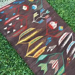 Puede incluir: Una alfombra marr&oacute;n con un patr&oacute;n geom&eacute;trico de hojas y flores estilizadas en tonos de rojo, amarillo, verde, azul y blanco. La alfombra tiene flecos en el borde superior.