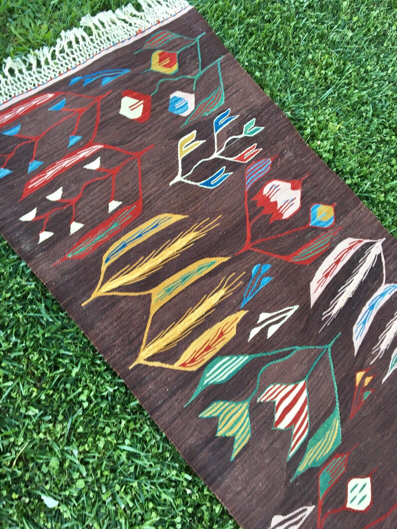 Puede incluir: Una alfombra marr&oacute;n con un patr&oacute;n geom&eacute;trico de hojas y flores estilizadas en tonos de rojo, amarillo, verde, azul y blanco. La alfombra tiene flecos en el borde superior.