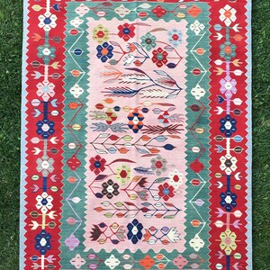 May include: A rectangular, hand-woven rug with a floral design. The rug features a central pink field with colorful floral motifs, framed by a green border and a red outer border. The rug has a fringe on the short ends.