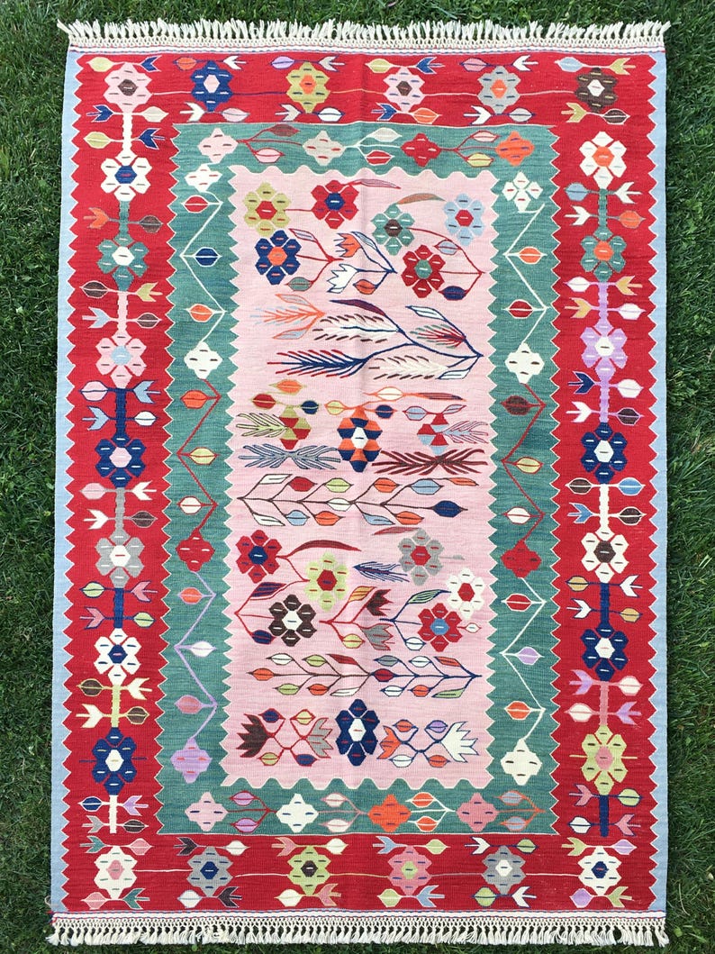 May include: A rectangular, hand-woven rug with a floral design. The rug features a central pink field with colorful floral motifs, framed by a green border and a red outer border. The rug has a fringe on the short ends.