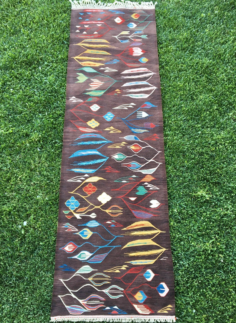 Puede incluir: Una alfombra larga y estrecha con fondo marr&oacute;n y un patr&oacute;n geom&eacute;trico colorido. El patr&oacute;n presenta una variedad de formas, incluyendo diamantes, hojas y espirales. La alfombra est&aacute; tejida con una variedad de colores, incluyendo rojo, azul, verde, amarillo y blanco.