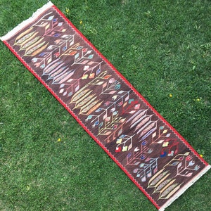 Puede incluir: Una alfombra de pasillo marr&oacute;n con un patr&oacute;n geom&eacute;trico de hojas y flores de colores. La alfombra tiene un borde rojo y flecos en los extremos.
